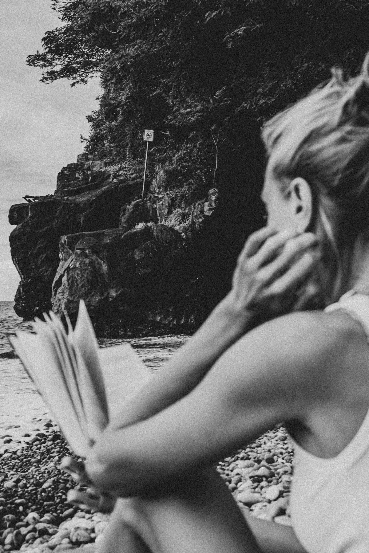 A reader with an open book on a pebbled beach beside rocky cliffs.